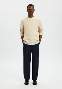 Beige stickad tröja med ribbade manschetter och nederkant, kombinerad med mörkblå vida byxor och svarta skor, framför en ljus och enkel bakgrund.
