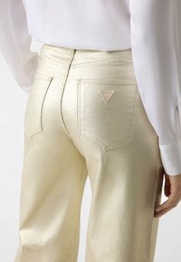Gouden metallic jeans met een gladde textuur, voorzien van achterzakken en een driehoekig logopatch. Gecombineerd met een witte blouse.