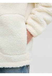 Biały, puszysty polar z kieszenią kangurzą i elastycznymi mankietami. Tekstura jest miękka z lekko podniesionym wzorem.