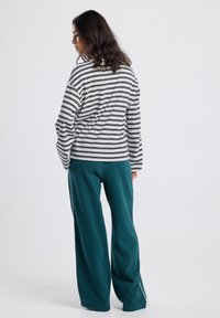 Woman with wavy hair wearing a black and white striped long-sleeve shirt and teal wide-leg pants, standing facing sideways against a white background.