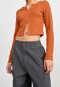 Top de manga larga recortado con acanalado en color naranja quemado, con cierre de cremallera. Combinado con pantalones grises de corte holgado que cuentan con bolsillos de carga.