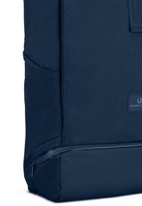 Donkerblauwe stoffen rugzak met zijvak, ondervak met ritssluiting en een klein rechthoekig logoplaatje aan de voorkant.