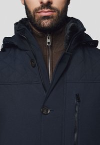 Granatowa pikowana kurtka z kapturem wyłożonym futerkiem, z brązową zapinaną na zamek warstwą wewnętrzną, zapięciem na guziki oraz wieloma kieszeniami zapinanymi na zamek.