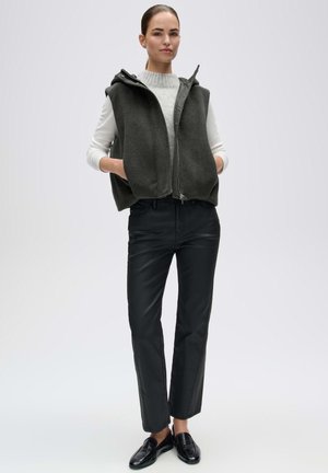 Donna che indossa un gilet senza maniche grigio scuro con zip sopra un maglione bianco, pantaloni neri a gamba dritta e mocassini neri, in piedi con le mani nelle tasche.