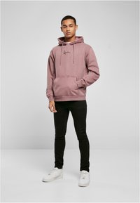 Mikina s kapucňou v tlmenej purpurovej farbe, z bavlneného zmesi, s klokankou a sťahovacími šnúrkami. Kombinovaná s čiernymi slim-fit nohavicami a bielymi teniskami.