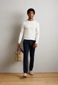Witte blouse met lange mouwen, marineblauwe broek, beige tas met bruine accenten, en lichte schoenen; model staat tegen een neutrale achtergrond.