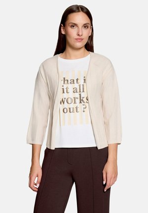 Woman wearing a beige open-front cardigan over a white shirt with gold text and dark brown pants, standing against a plain background.