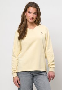 Jonge vrouw met lang bruin haar, gekleed in een lichtgele V-hals trui en lichtgrijze spijkerbroek, staand tegen een eenvoudige lichte achtergrond.