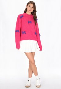 Flauschiger rosa Pullover mit lila Schleifenakzenten, Rundhalsausschnitt, langen Ärmeln und lockerer Passform. Kombiniert mit einem weißen, plissierten Rock und Turnschuhen.