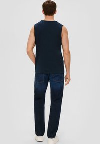 Marineblaues ärmelloses Shirt und dunkelblaue Jeans in klassischer Passform mit Gesäßtaschen und glatter Textur. Die Schuhe sind weiß und im lässigen Stil.