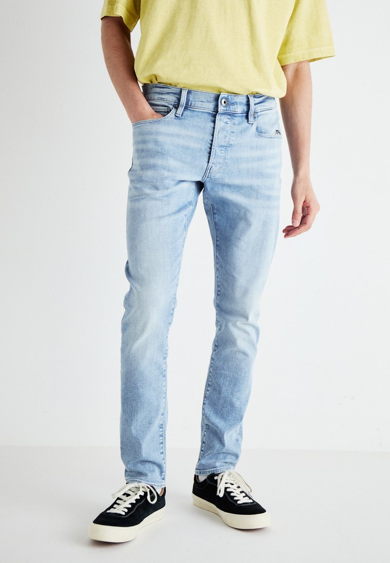 Ljusblå denimjeans med slim fit, fem fickor, dragkedja och en lätt urtvättad finish, i kombination med svarta sneakers och en gul t-shirt.