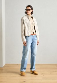 Giacca cropped beige sopra una camicia button-up beige, jeans dritti azzurri chiari e sneakers gialle con dettagli marroni, occhiali da sole indossati.