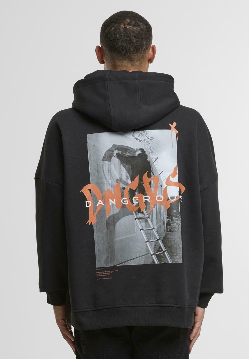 Hoodie surdimensionné noir avec un imprimé graphique dans le dos, affichant le mot "DANGEREUX" en orange et une image monochrome au-dessus.