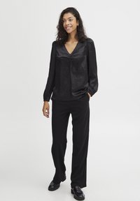 Schwarze Satinbluse mit V-Ausschnitt und gerafftem Detail an der Vorderseite, kombiniert mit schwarzen weit geschnittenen Hosen und klobigen schwarzen Schuhen.