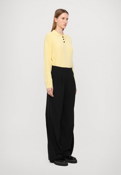 Camisa polo amarilla de manga larga con cuello y tres botones negros, combinada con pantalones de tiro alto de pierna ancha negros y zapatos negros gruesos.