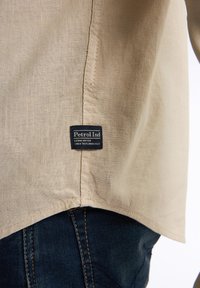 Beige shirt van lichtgewicht stof, met een gekromde zoom en een zwart label met witte tekst dat aan de onderrand is genaaid. Donker denim zichtbaar eronder.