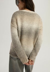 Sweter robiony na drutach w beżowo-szarym ombre, charakteryzujący się luźnym krojem, długimi rękawami oraz strukturalnym splotem. Widok z tyłu podkreśla miękkość swetra.