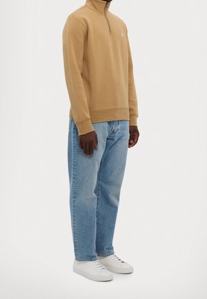 Mann trägt ein sandfarbenes Sweatshirt mit Viertel-Reißverschluss, hellblaue Jeans und weiße Turnschuhe, steht vor einem schlichten weißen Hintergrund.