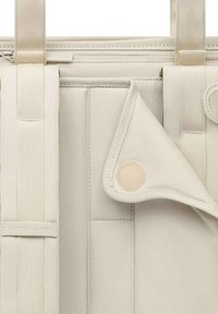 Sac en tissu beige avec des sangles réglables et un rabat plié fixé par un bouton-pression rond beige, montrant les détails de la texture et des coutures.