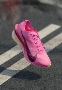 Løpesko med en lys rosa overdel, teksturert materiale, buet form, en fremtredende lilla swoosh, og en gul yttersåle.