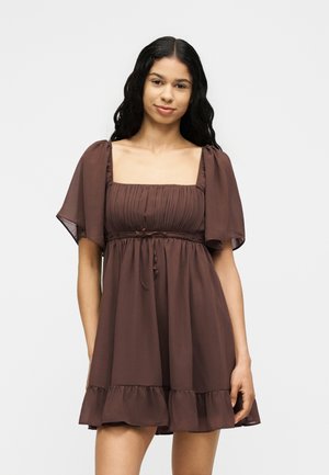 Ung kvinde med langt sort bølget hår iført en kort, løs brun kjole med pufærmer og en flæsekant, stående mod en ensfarvet baggrund.