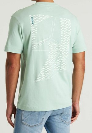 Homme portant une chemise vert clair avec un motif géométrique blanc abstrait et un texte "CHASIN'" dans le dos, assortie à un jean bleu.