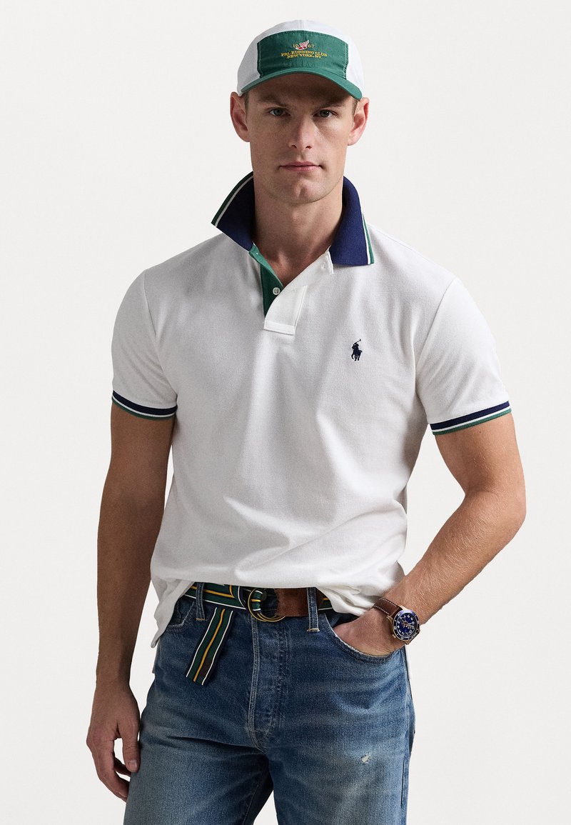 Vit polo skjorta med marinblå krage och gröna detaljer; kombinerad med blåjeans och ett mönstrat bälte. Kapsylen har gröna och vita färger med logotyp.