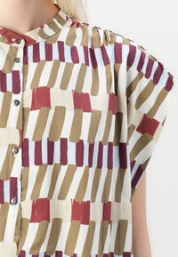 Patterned blouse with a stand collar, featuring a brushstroke design in red, brown, and light blue on a cream background, with button closure.