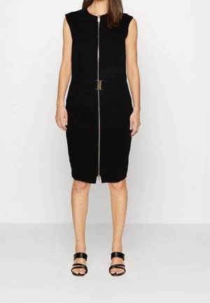 Femme portant une robe noire sans manches longueur genoux avec une fermeture éclair à l'avant et une ceinture à la taille, associée à des sandales noires à bout ouvert.