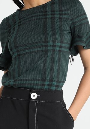 Žena v zeleno-černém kostkovaném topu s krátkým rukávem zastrčeném do černých kalhot s bílým knoflíkem a šitím, s rukou v kapse.