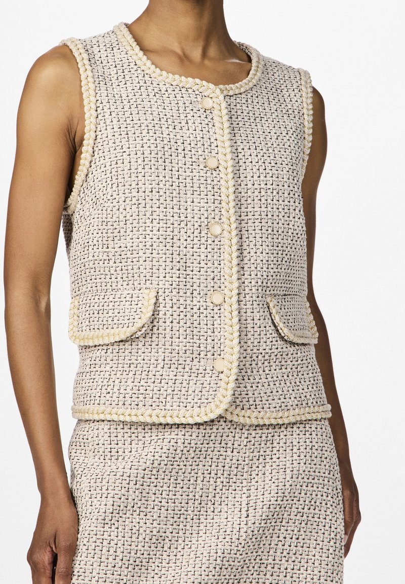 Kamizelka bez rękawów w kolorze kremowym z teksturowanym splotem. Posiada dwie przednie kieszenie, plecioną lamówkę oraz zapięcie na guziki.