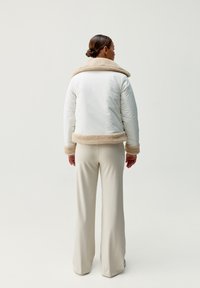 Biała izolowana kurtka z brązowym kołnierzem i brzegiem z owczej skóry. W zestawieniu z kremowymi spodniami w kształcie dzwonów. Minimalistyczny design na jasnym tle.