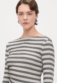 Mujer joven con cabello corto castaño, llevando pendientes de aro de plata y una camiseta de manga larga de rayas en blanco y negro, con un fondo neutro.