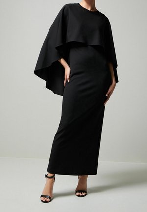 Femme portant une robe noire moulante longue avec un voile type cape et des talons noirs à bride cheville, posant devant un fond uni.