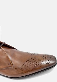 Zapato de vestir de cuero marrón con punta afilada. Presenta detalles perforados en la parte superior y una textura suave. Diseño con cordones.