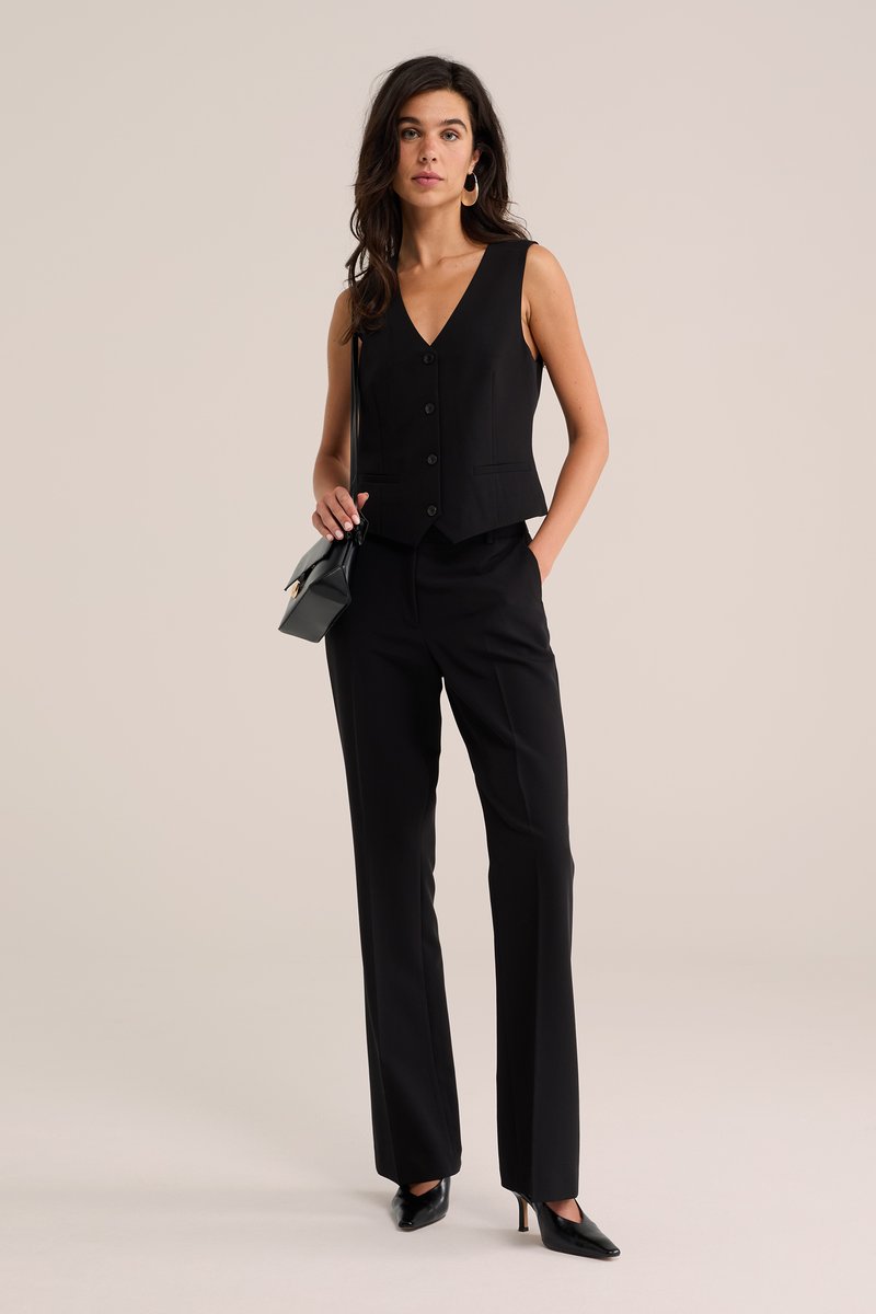 Mujer con chaleco negro sin mangas abotonado, pantalones negros, tacones negros, llevando un pequeño bolso negro de hombro, de pie frente a un fondo liso.