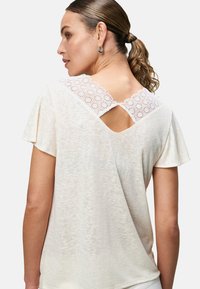 Lichtbeige top met korte mouwen, voorzien van een decoratieve witte kanten accenten op de rug, met een sleutelgatenontwerp boven de schouderbladen.