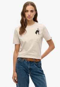 Camiseta de algodón color beige claro con un gráfico de un gato negro en el bolsillo del pecho. Manga corta y corte relajado, combinada con jeans azules.