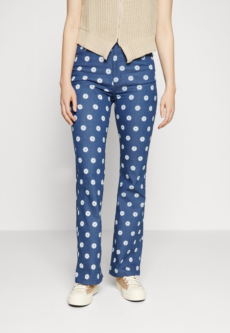 Calças à boca de sino em denim azul escuro com estampado de margaridas brancas; apresenta um cós e bolsos; combinadas com um top de malha bege claro e ténis.