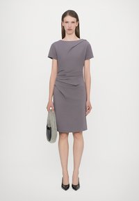 Gray fitted dress with short sleeves, draped detailing at the waist, paired with a black handbag and black pointed heels.