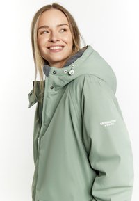 Lyst grøn vandtæt jakke med hætte, grå foer, lynlås lukning og logodetalje på ærmet. Har trykknapper og lommer.