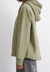 Olivgrön hoodie i mjukt material med en känguruficka, ribbade muddar och en huva med snörning, visad från sidan.