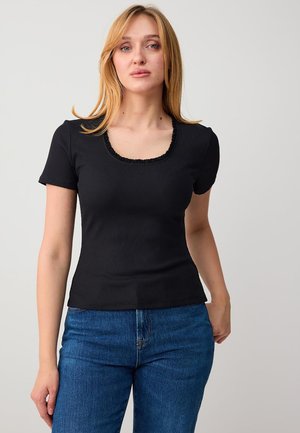 Jonge vrouw met blond haar, gekleed in een zwart topje met korte mouwen en een vierkante halslijn, en een blauwe spijkerbroek, tegen een effen achtergrond.