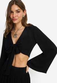 Μαύρο cropped τοπ με μακριά καμπανούλα μανίκια, κόμπο μπροστά και βαθύ V λαιμόκοψη· συνδυασμένο με μαύρη φούστα με ελαστική μέση.