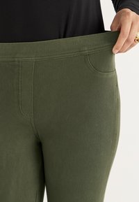 Olijfgroene broek met een brede elastische tailleband en voorzakken, gemaakt van een gestructureerde, rekbare stof. Close-up van het taillergebied.