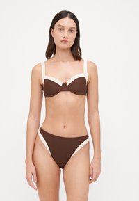 Brūns bikini komplekts ar krēmkrāsas akcentiem. Topam ir strukturēts dizains ar platām siksnām un centrālo aizdari. Apakšušķirnes ir ar augstiem izgriezumiem.