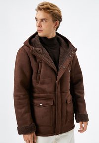 Brun, faux suede jakke med hætte, blød pelsforing, frontlommer og knapdetaljer. Har en tekstureret overflade og fyldige manchetter.