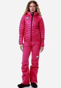 Giacca imbottita rosa con cappuccio presenta un design trapuntato e cerniera nera. I pantaloni da sci rosa abbinati hanno dettagli con logo, creando un look coerente.