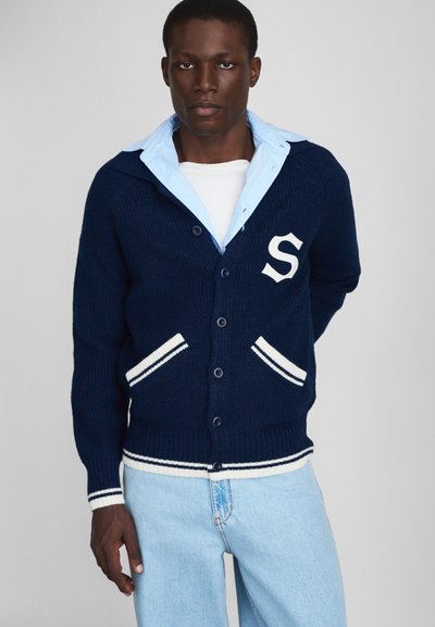 Cardigan en tricot marine avec des finitions blanches, arborant une lettre « S » sur la poitrine, fermeture à bouton et deux poches avant. Chemise à col bleu clair en dessous.