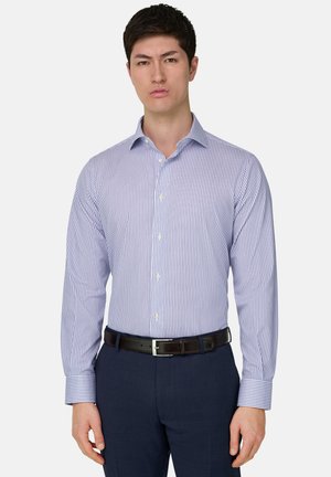 Mann trägt ein blau-weiß vertikal gestreiftes Hemd, das in marineblaue Hosen mit schwarzem Gürtel gesteckt ist, steht vor einem schlichten Hintergrund.